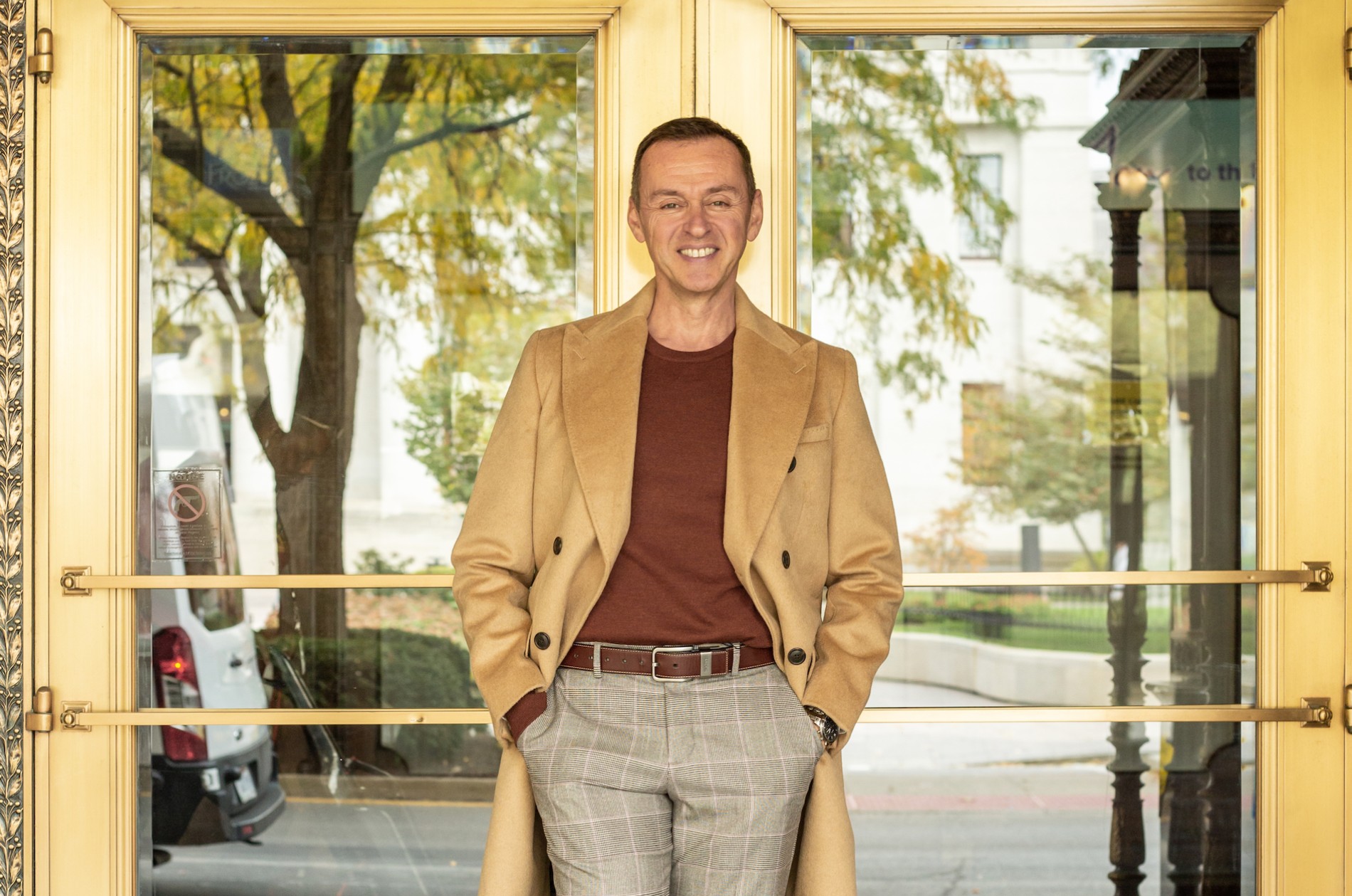 Person dressed in business casual in autumnal colors standing on a tile floor beside old brass doors with windows to a tree-lined street.