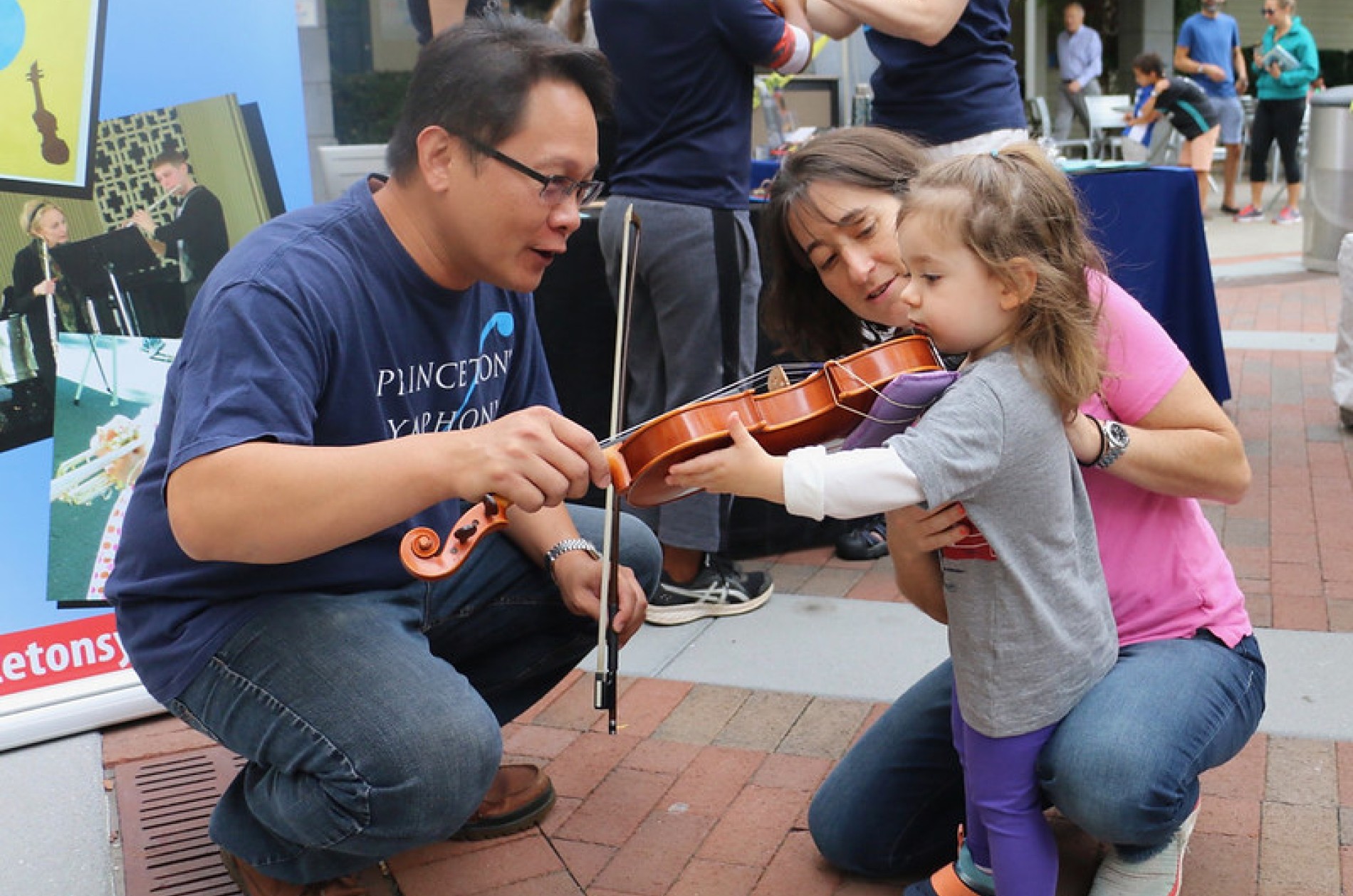 Musician squatting down to help small child with accompanying parent play the violin at an outdoor event.