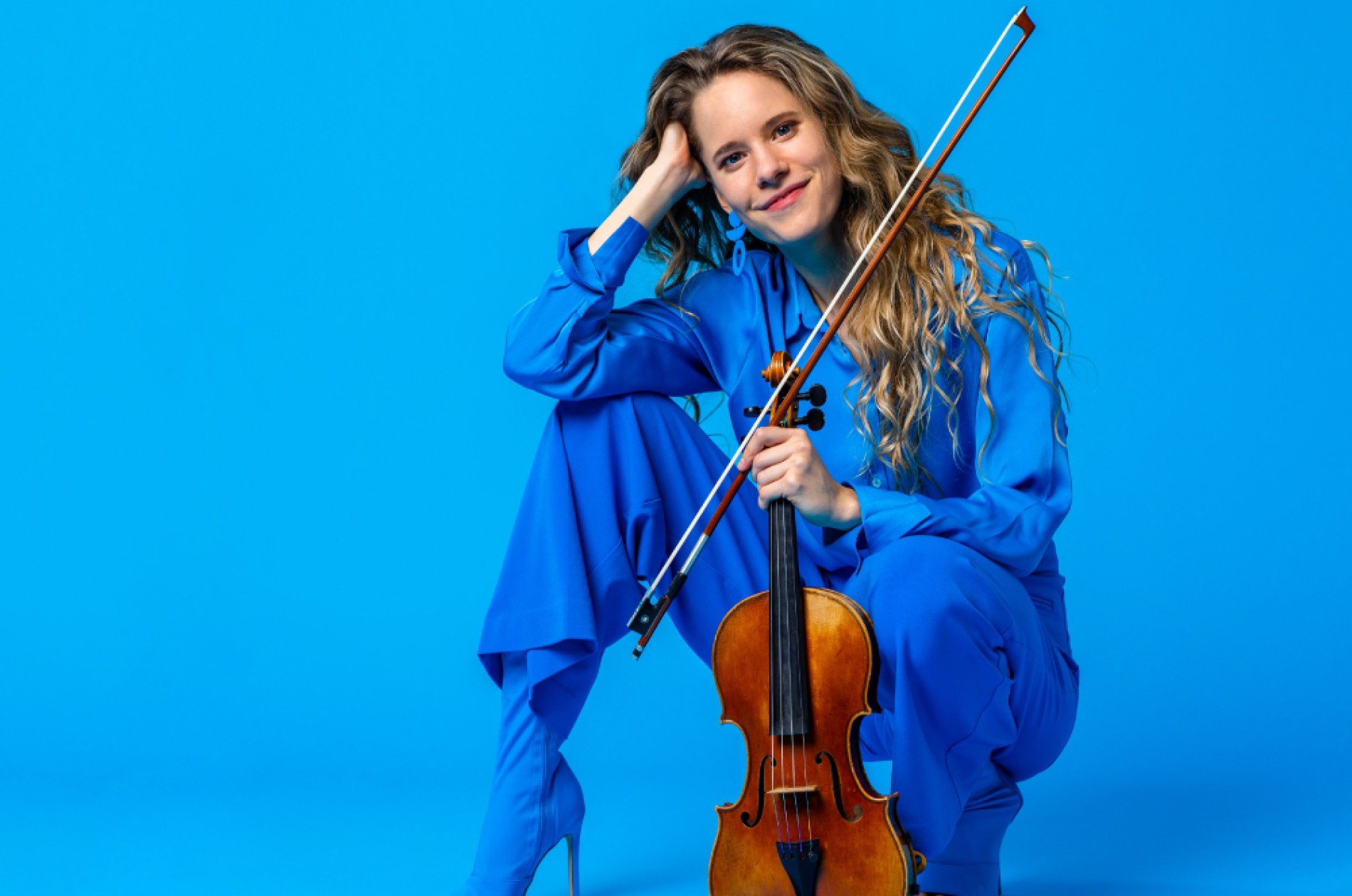 A young woman smiles and squats while holding her violin. 