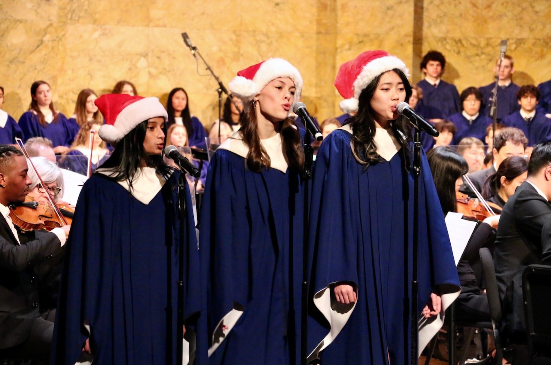 Three choir members in blue choir robes and red Santa hats singing on stage as a trio with a few orchestra members and the full choir in the background.