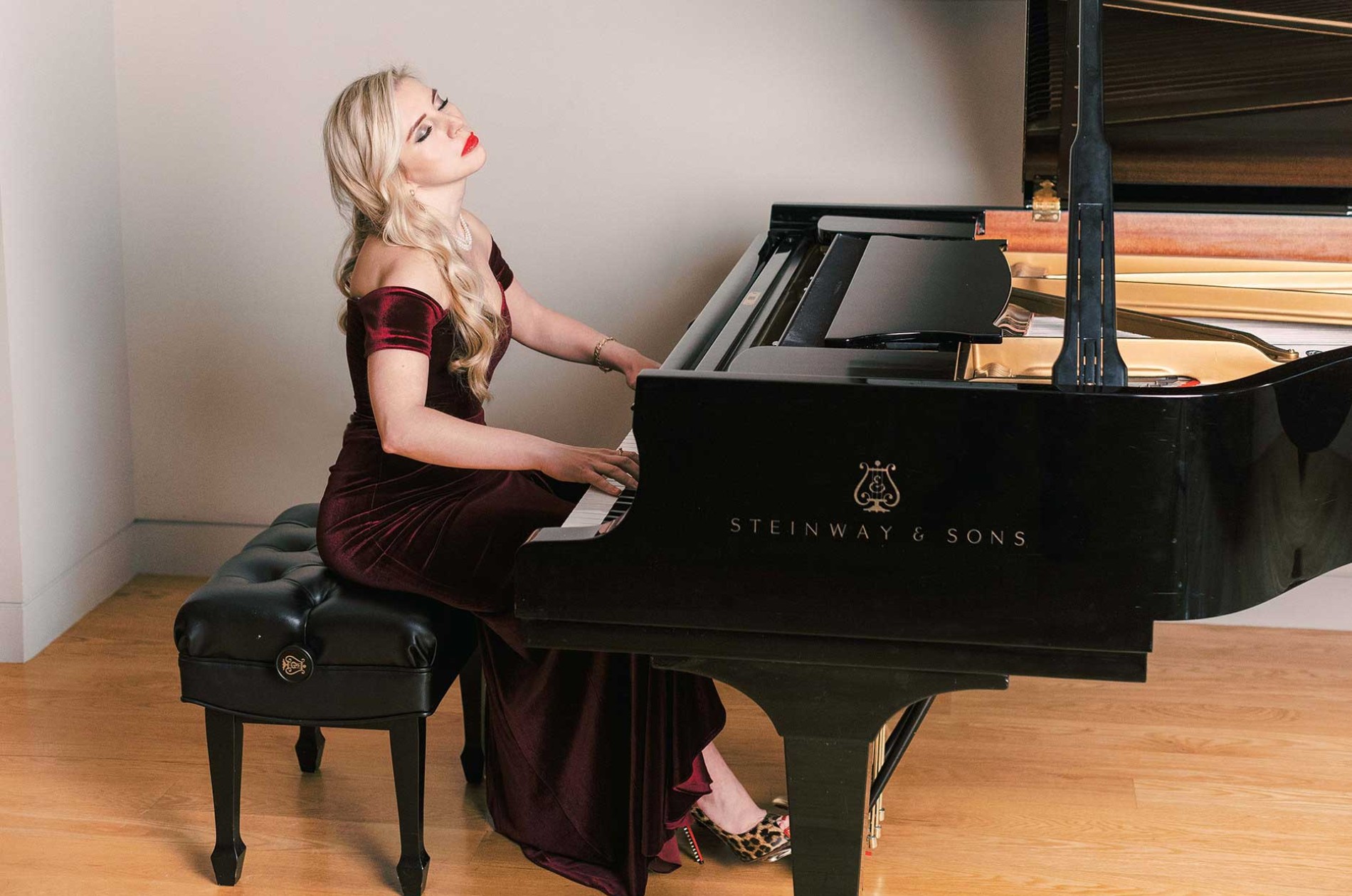 Women in a red velvet gown playing music on a grand piano upon a light wood floor.