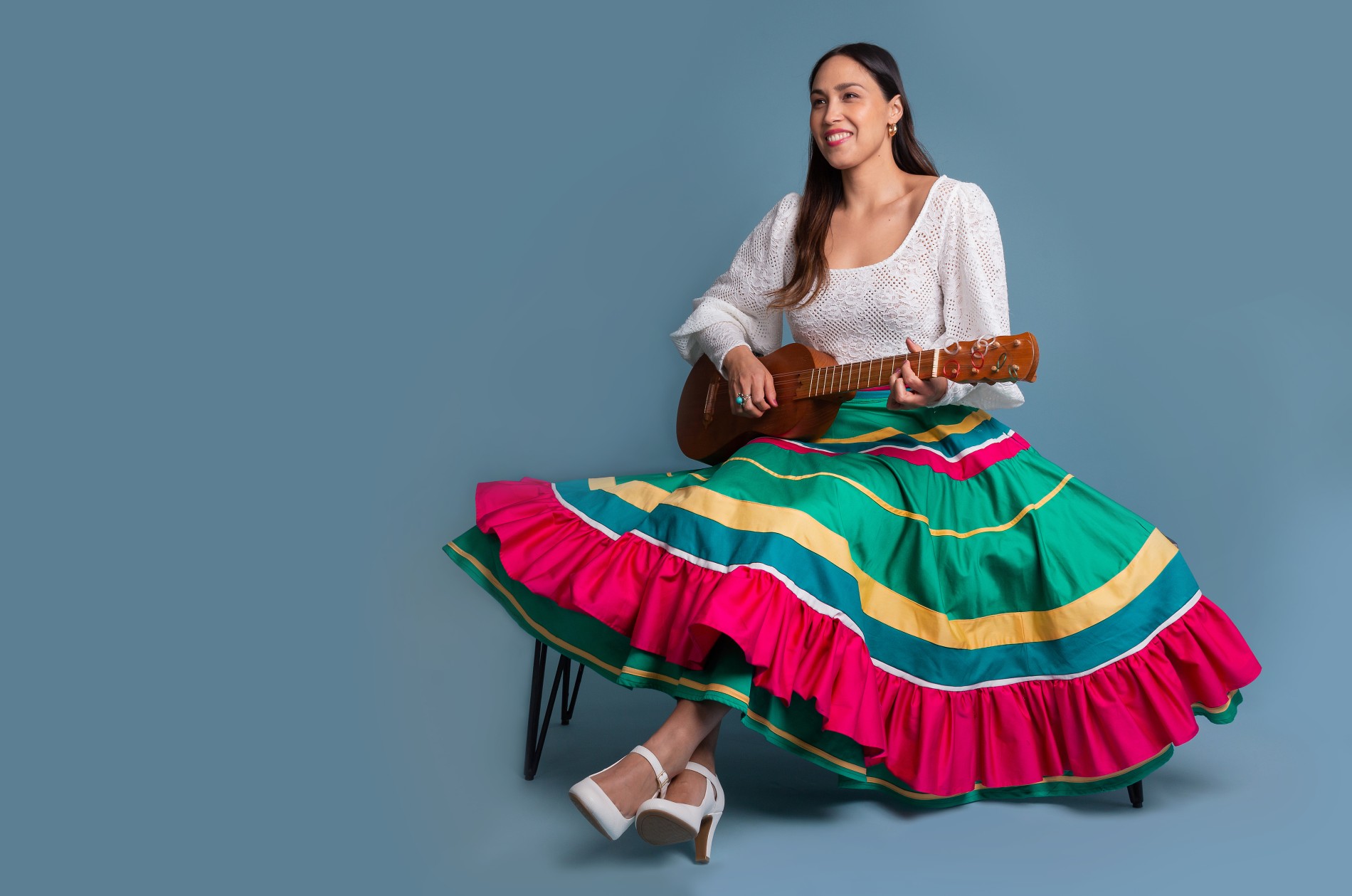 Sonia de los Santos, seated on a bench and wearing a colorful, full skirt and white blouse, strumming a wooden guitar.