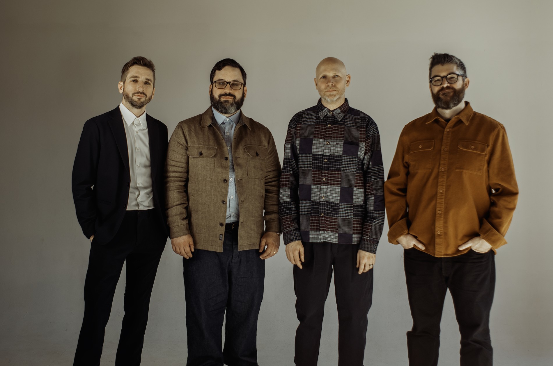 Sō Percussion group shot - Four men standing in casual clothes against a greyish backdrop
