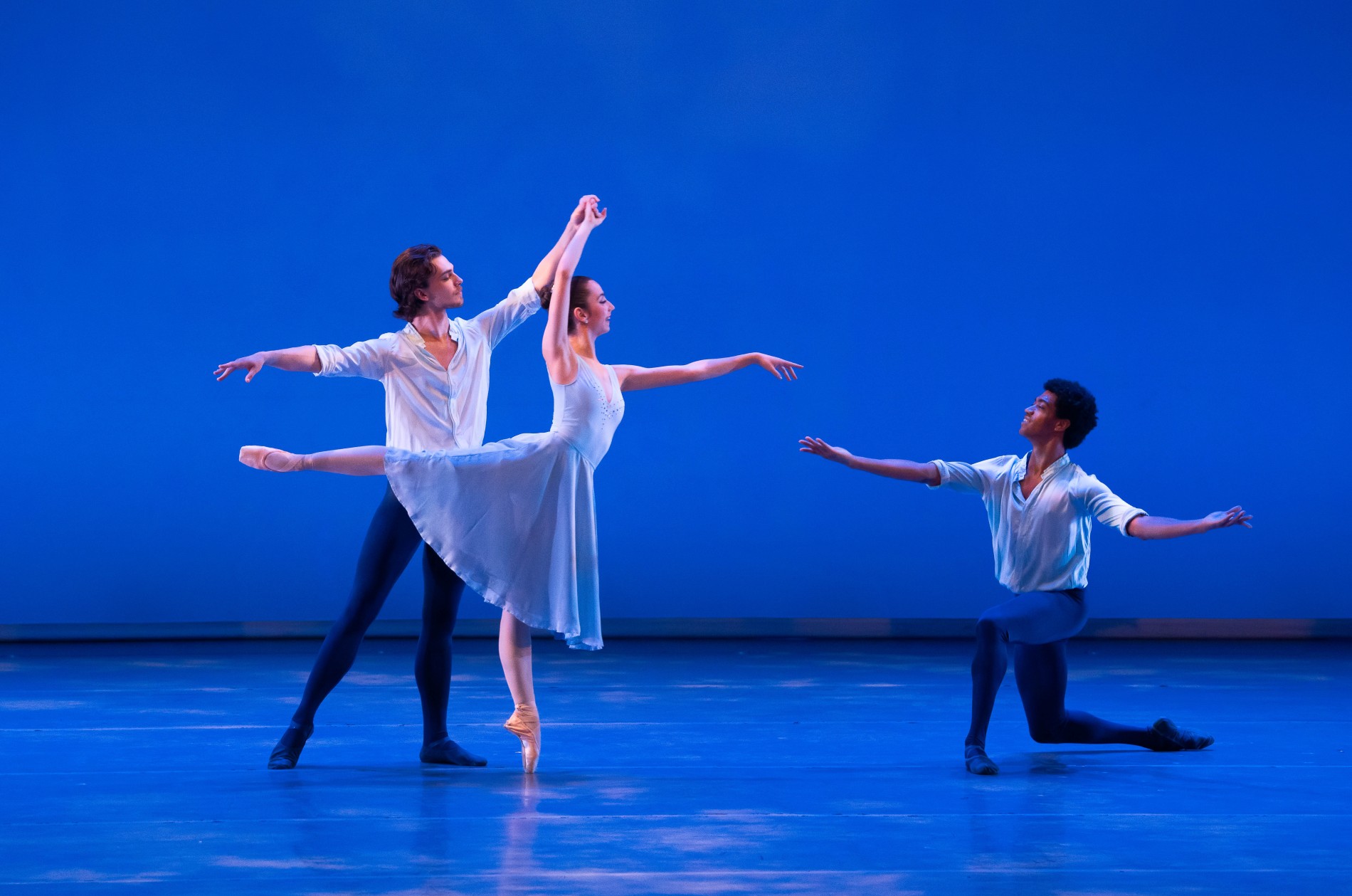 Three ballet dancers performing on stand; the one in center performing an arabesque with the two male dancers in black tights and coordinating shirts pose standing and kneeling with arms spread open wide.